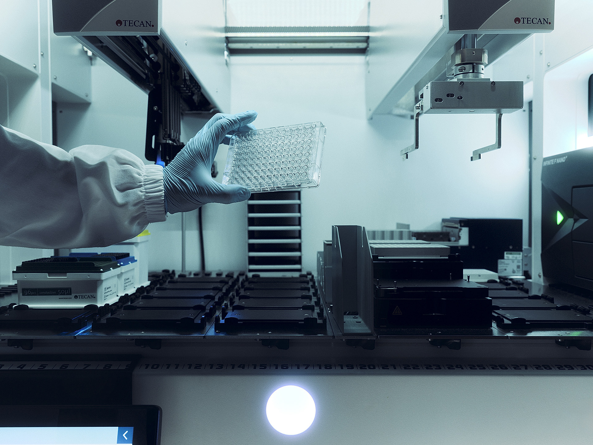 Una mano guantata posa una micropiastra su una postazione di automazione da laboratorio: biologia e ingegneria in un solo gesto.